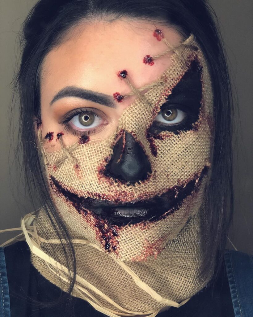 A person wearing scarecrow makeup, with burlap-like material stitched to the face, black hollow eyes, and a wide, bloody grin.