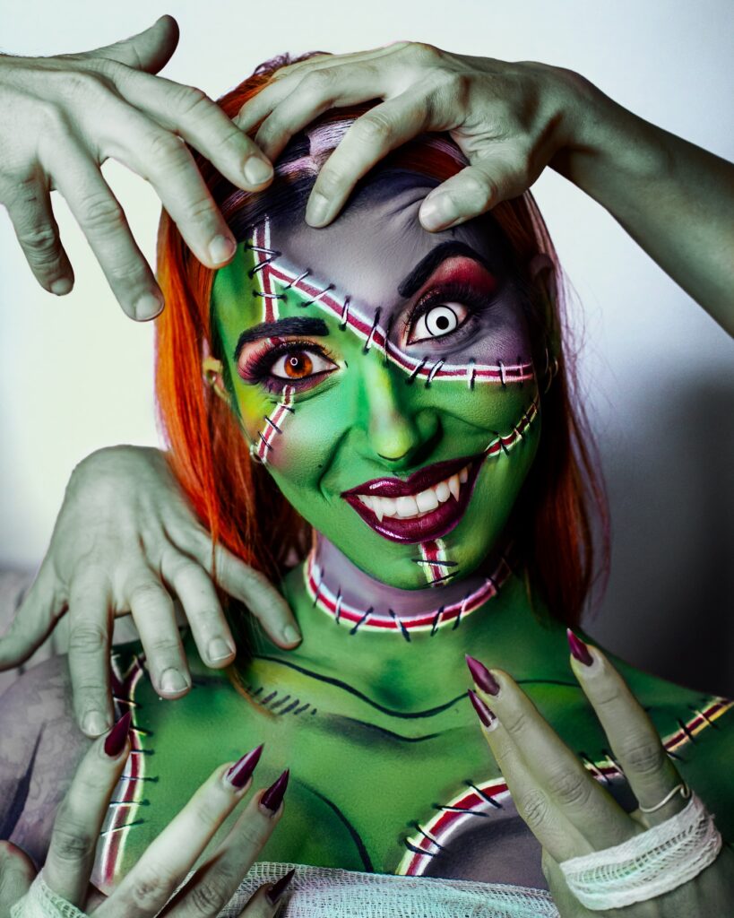 Person wearing Frankenstein-inspired Halloween makeup with green and grey stitched patches, white contacts, bold eye makeup, red lips, and surrounded by multiple eerie hands.