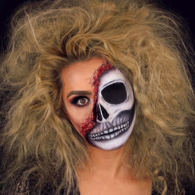 Person wearing half-glam, half-skeleton Halloween makeup with one side featuring bold makeup and the other side a skull with exposed bloody details, complemented by wild, teased hair.