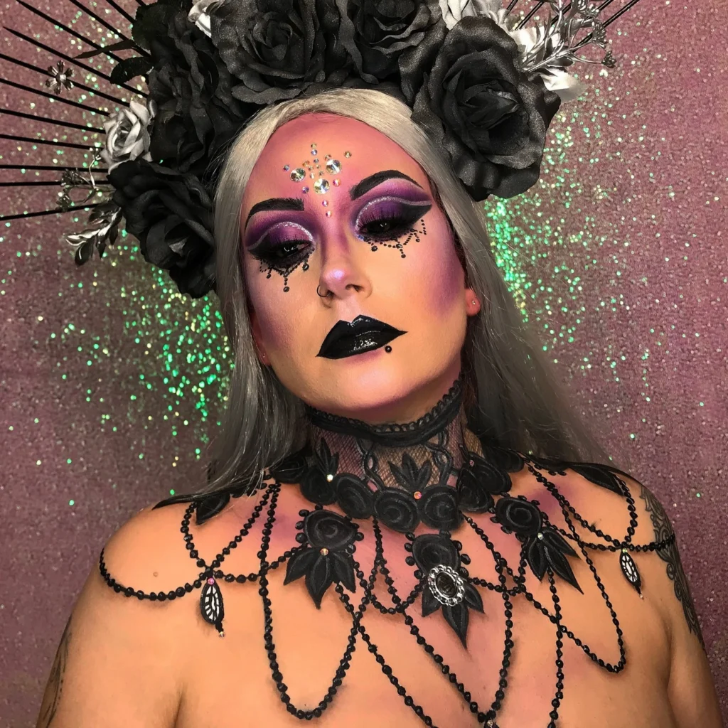 Close-up of a woman with Gothic glam Halloween makeup, featuring dark purple eyeshadow, black lipstick, elaborate face jewels, and a black lace choker with black roses. She wears a large black floral headdress and a silver wig.