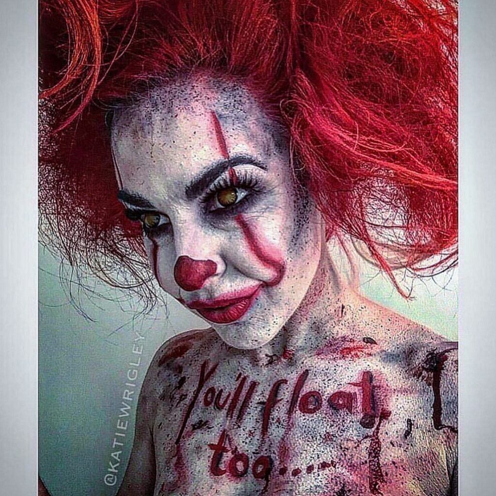 "Person wearing Pennywise-inspired Halloween makeup with red facial markings, a red nose, wild red hair, and the phrase 'You'll float too' painted across the chest.