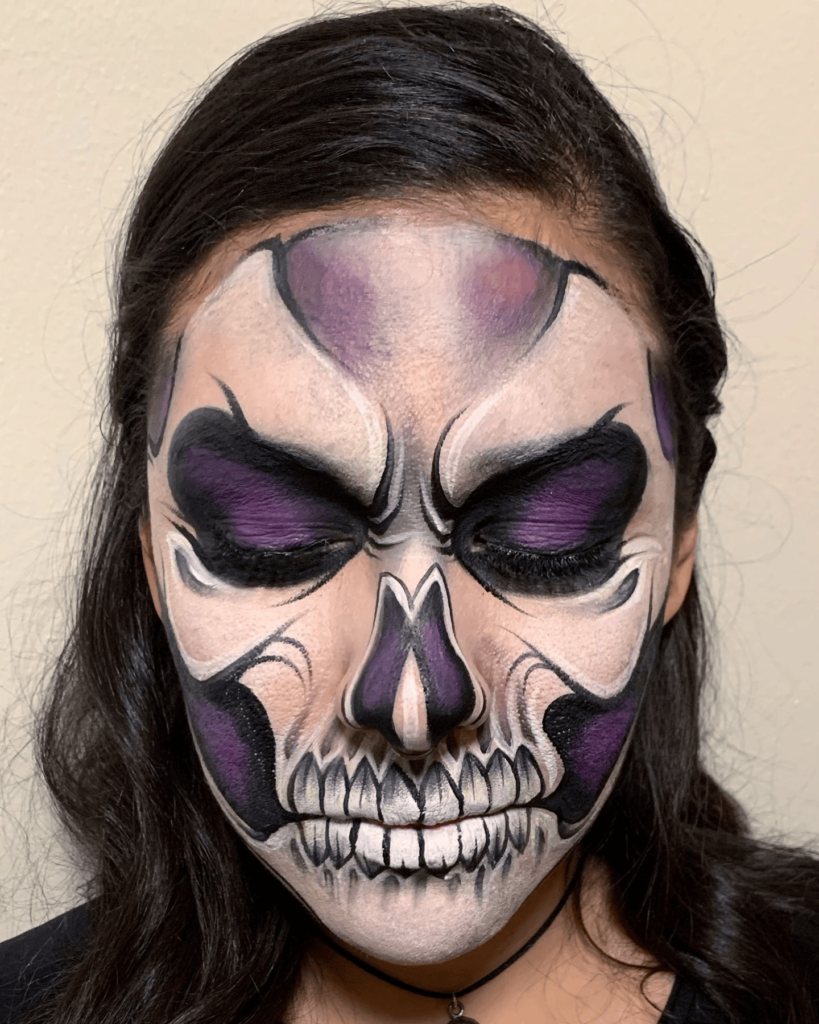 A person with purple and black skull face paint, featuring hollowed eyes and cheekbones with intricate skeletal details.