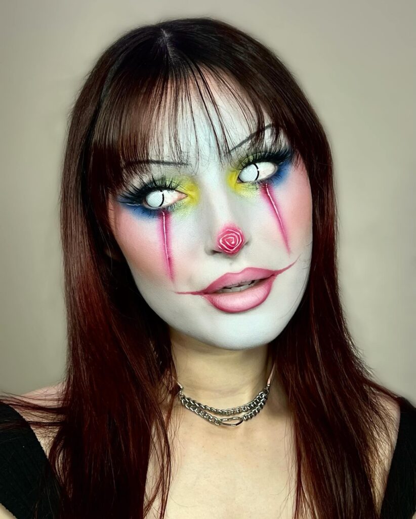 Woman with colorful pastel clown makeup featuring bright eyeshadow, a glowing rose-shaped nose, and subtle clown lip details. She wears a silver chain necklace, adding a modern touch to the clown-inspired appearance.