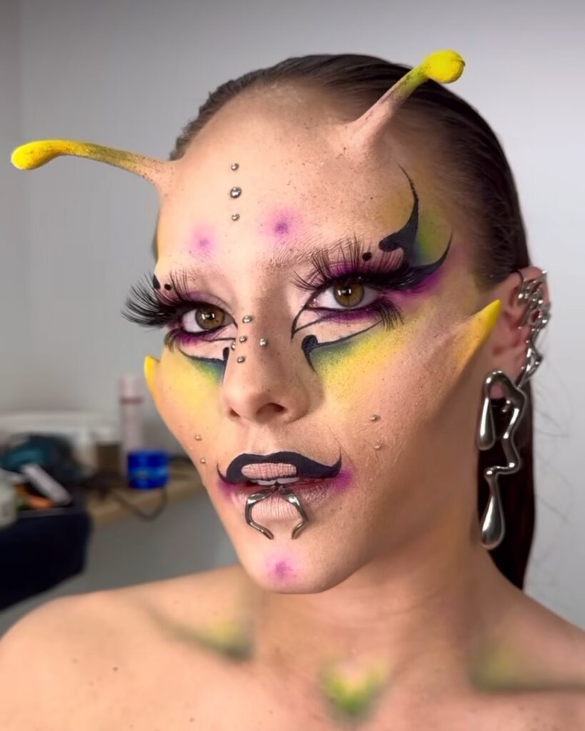 Person wearing alien-inspired makeup with vibrant yellow, purple, and black colors on the face, featuring antennae-like structures on the forehead, dotted embellishments, and metallic piercings for a futuristic, extraterrestrial look.