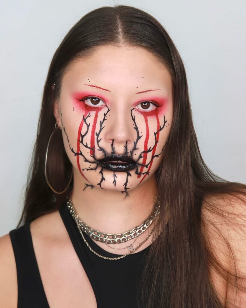 Person with Halloween makeup featuring black and red vein designs from the eyes to the mouth, paired with bold black lipstick and red eyeshadow for a vampiric gothic appearance.