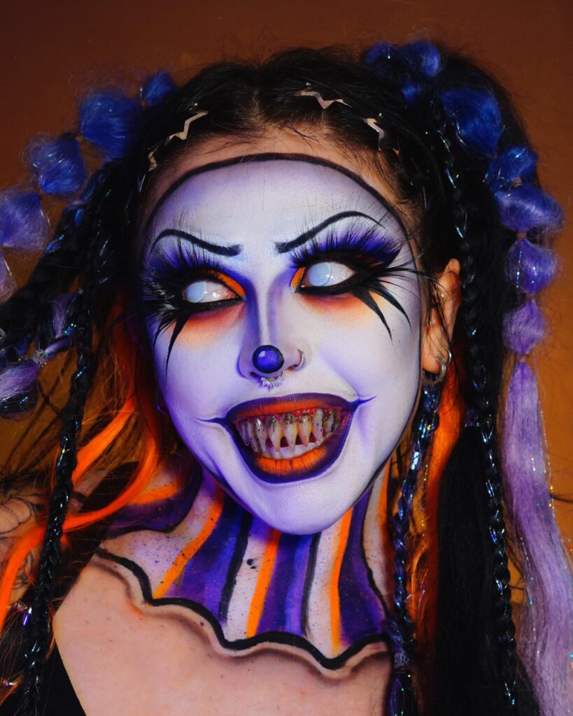 Person wearing creepy clown-inspired Halloween makeup with sharp, discolored teeth, glowing white eyes, and bold orange and purple accents, styled with colorful braids and a sinister grin.