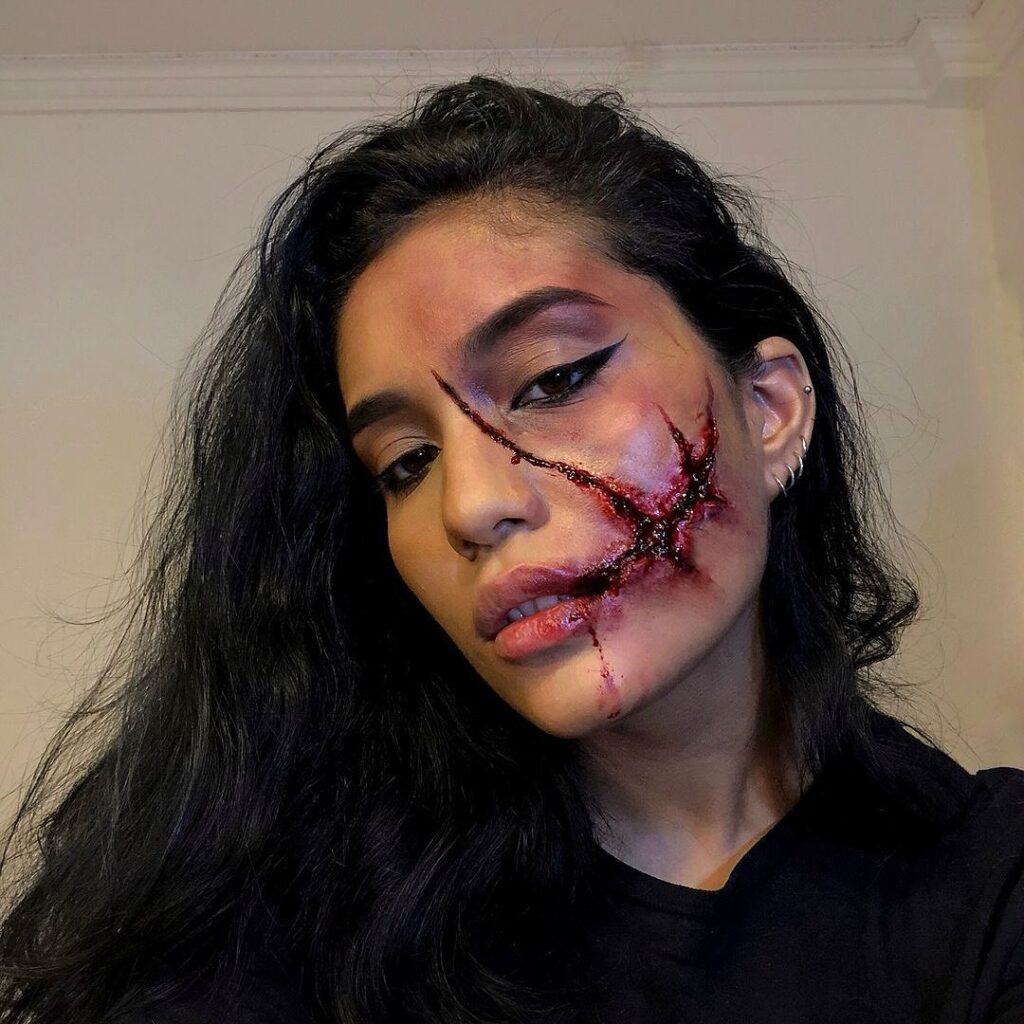 Person wearing realistic scar Halloween makeup with a deep, bloody wound across the cheek, paired with natural eyeliner and tousled hair for a dramatic look.