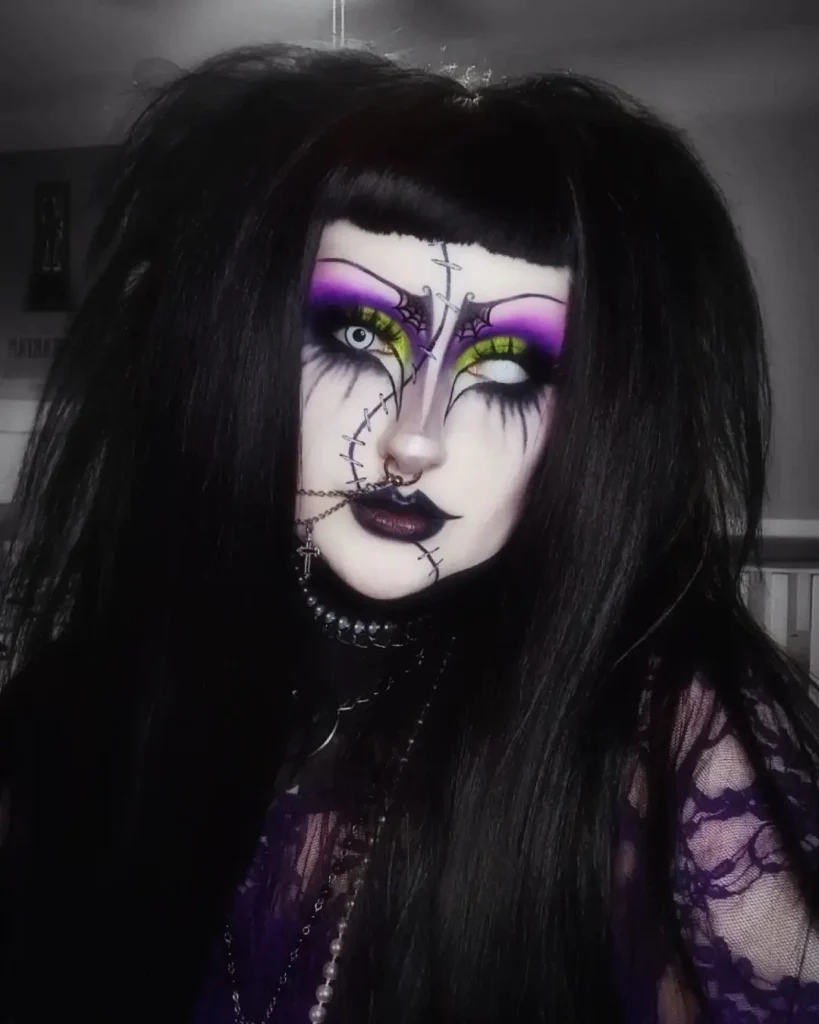 Person wearing stitched gothic Halloween makeup with purple and neon green eyeshadow, black lipstick, and voluminous black hair, styled with stitched scars across the face.