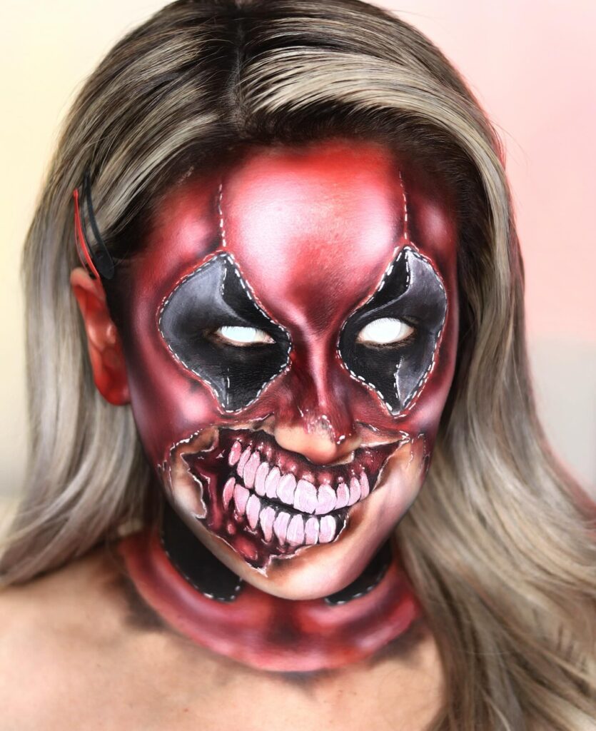 Person wearing terrifying monster Halloween makeup with exposed teeth, red and black skin, white eyes, stitched mask details, and a wide grotesque grin for a chilling and horrifying look.