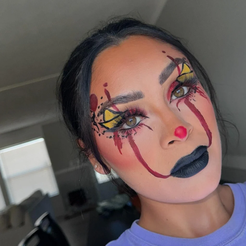 Close-up of a woman with Halloween makeup featuring a clown-inspired design with bold yellow and red eye makeup, a red nose, dramatic black lips, and smeared red lines under the eyes, creating a playful yet eerie appearance.