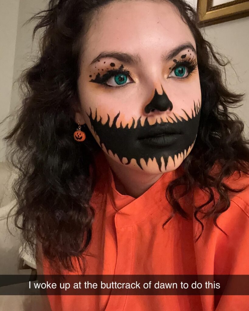 A person wearing a Jack-O'-Lantern themed makeup featuring black flame-like details around the mouth and nose, with bright teal eyes and pumpkin earrings for a festive touch.