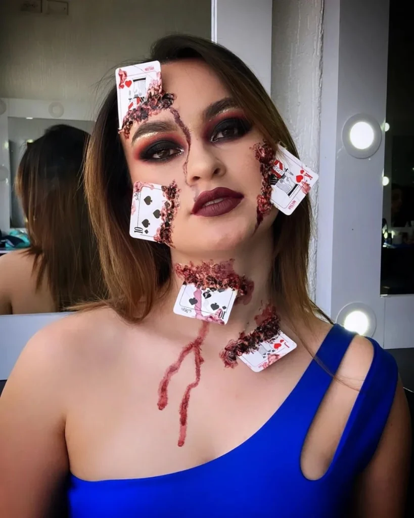 Close-up of a woman with Halloween makeup featuring playing cards embedded into her face and neck, with realistic bloody effects and dark, smoky eye makeup. The look is both eerie and detailed, creating a striking horror-inspired effect.