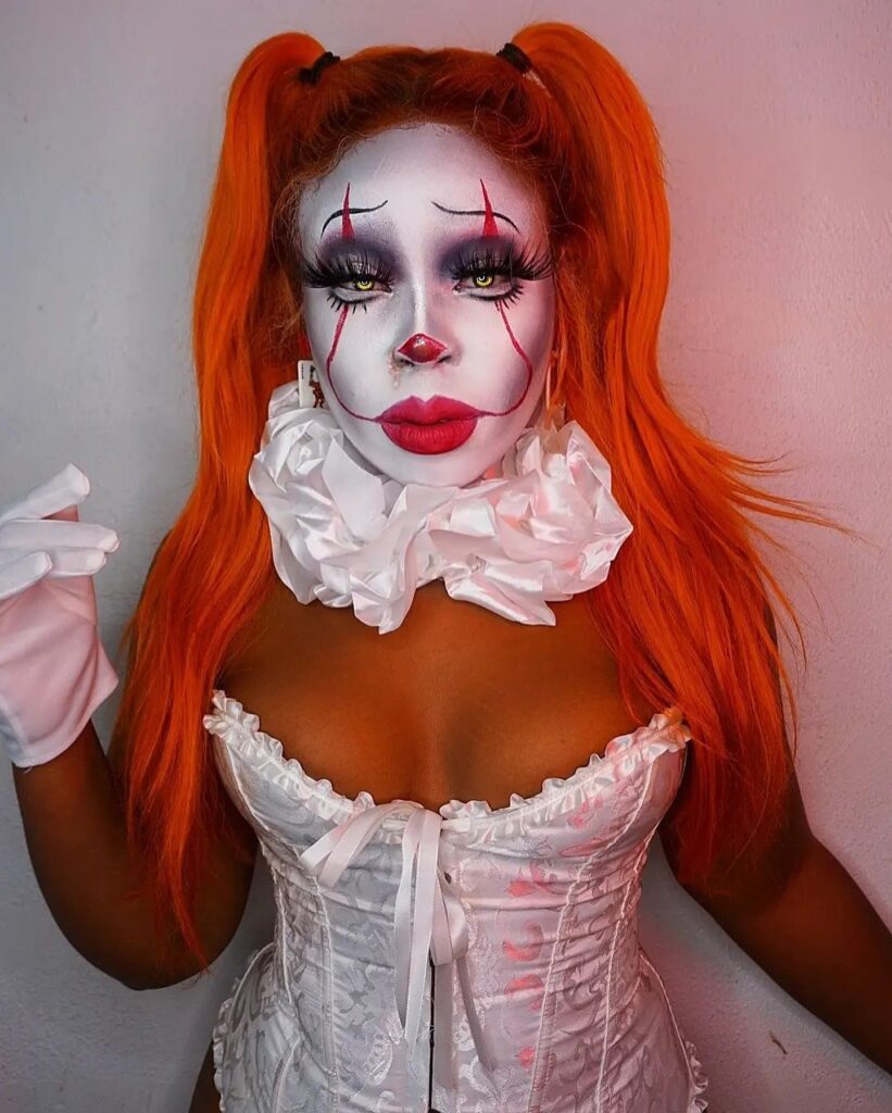 A person dressed as a glamorous Pennywise, with a white face, red makeup details, orange pigtails, and a white corset with a ruffled collar.