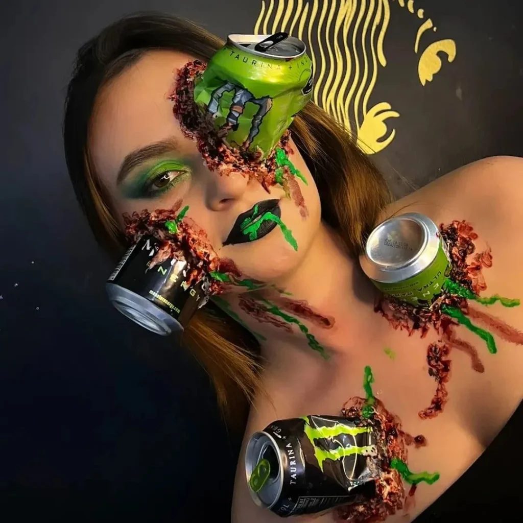 Close-up of a woman with Halloween makeup featuring crushed Monster Energy cans embedded into her face and body, with realistic bloody effects and vibrant green makeup accents. The look is both shocking and highly detailed, creating a striking horror-inspired effect.