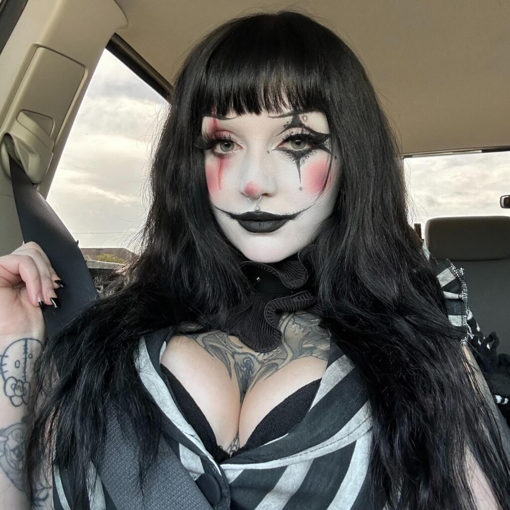 Woman with gothic clown makeup, featuring dark lips, black and white makeup with pink blush on cheeks, and dramatic eyes, sitting in a car.