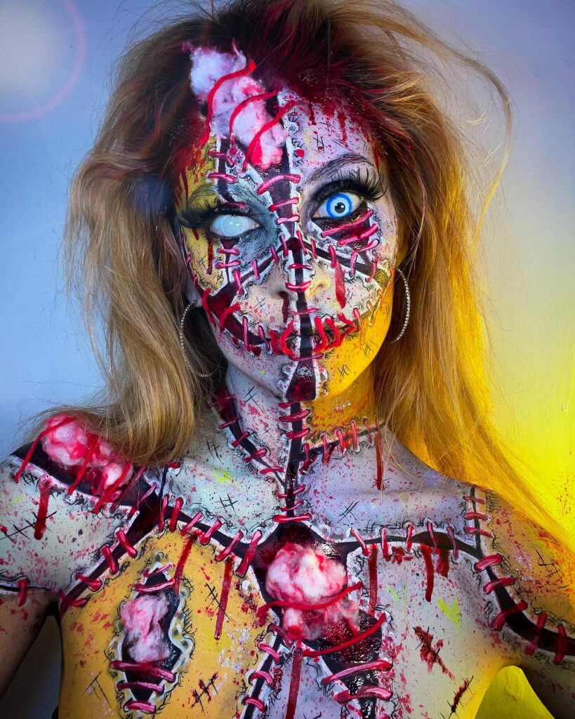 Person wearing stitched horror doll Halloween makeup with exposed wounds, red stitches, dramatic lashes, and white contacts for a gruesome look.