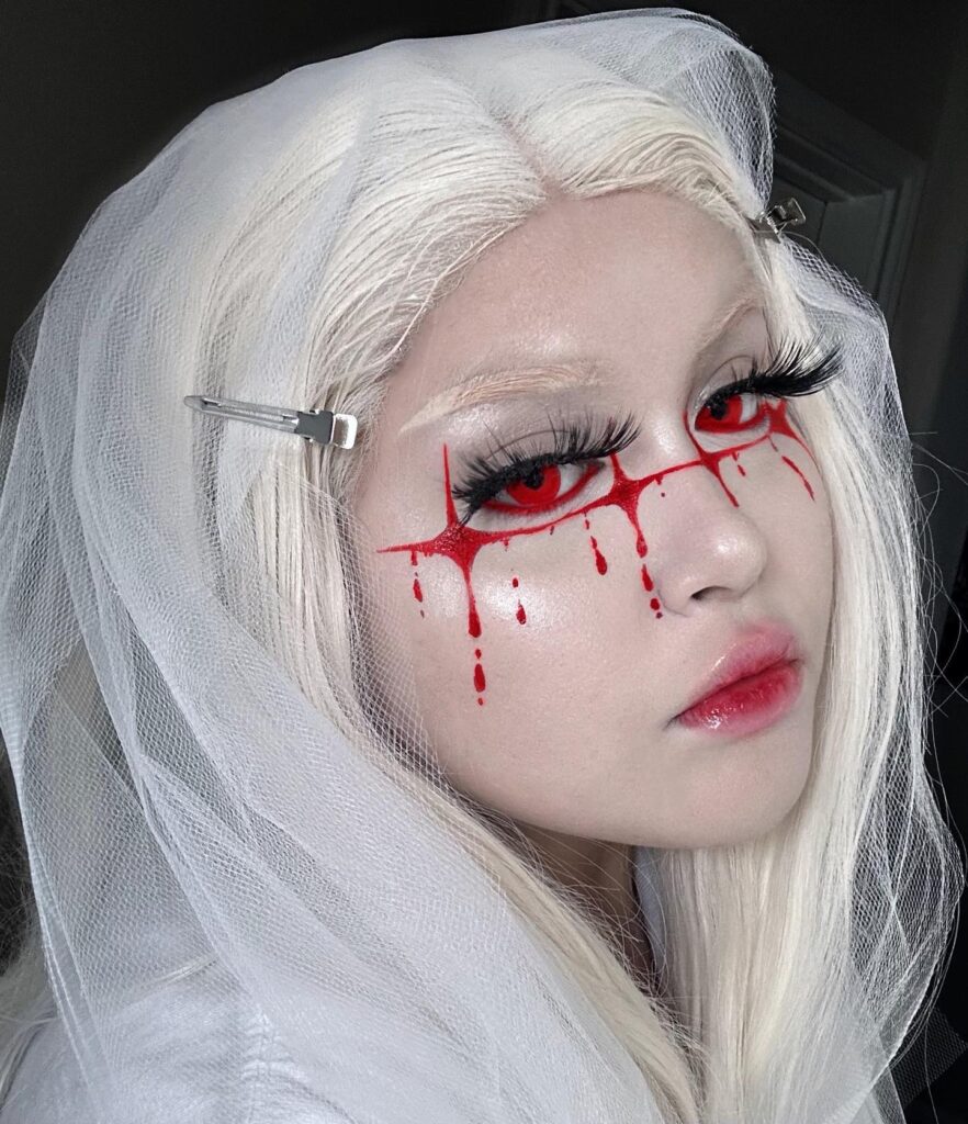 Person wearing ghost bride-inspired Halloween makeup with red dripping eye accents, long lashes, glossy lips, and a white veil for a haunting look.