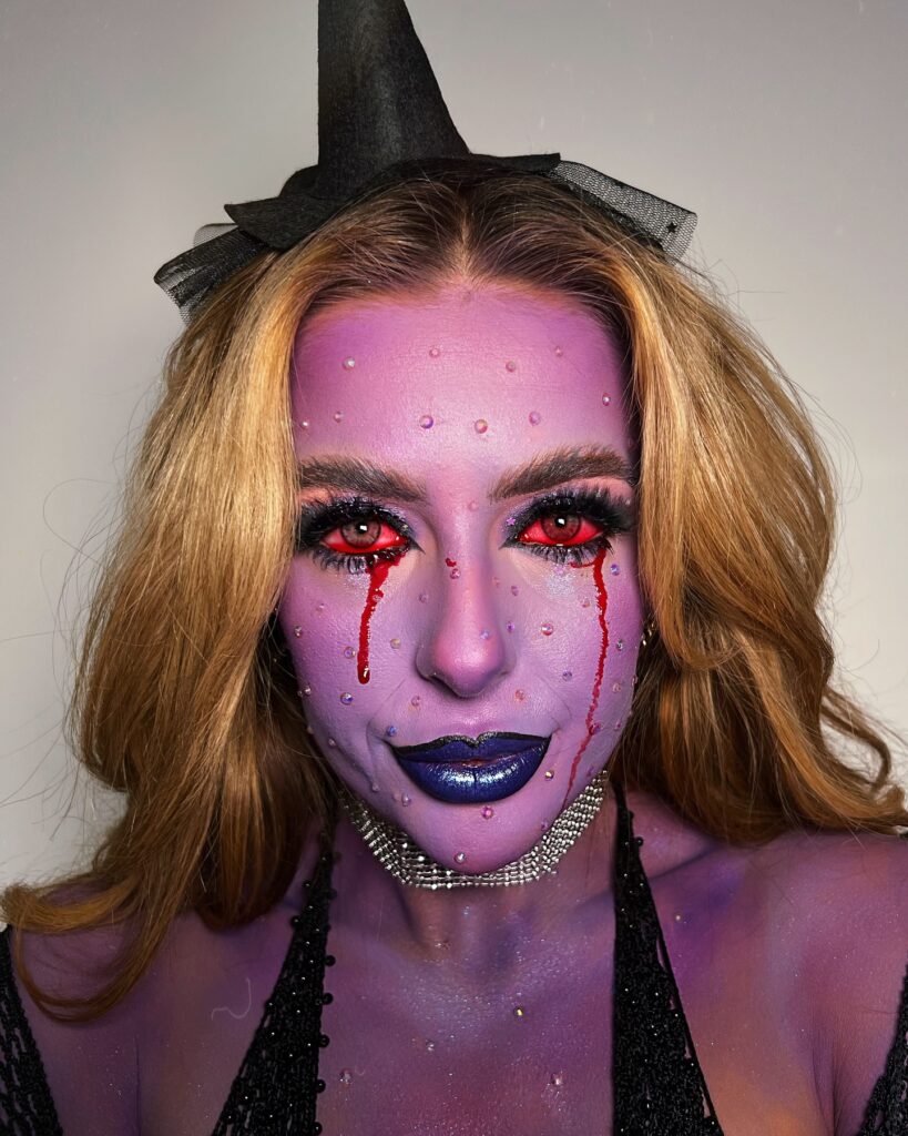 Person wearing an enchanted witch-themed Halloween makeup with a purple complexion, glitter accents, red eyes, glossy blue lips, blood tears, and a black witch hat for a magical and spooky look.