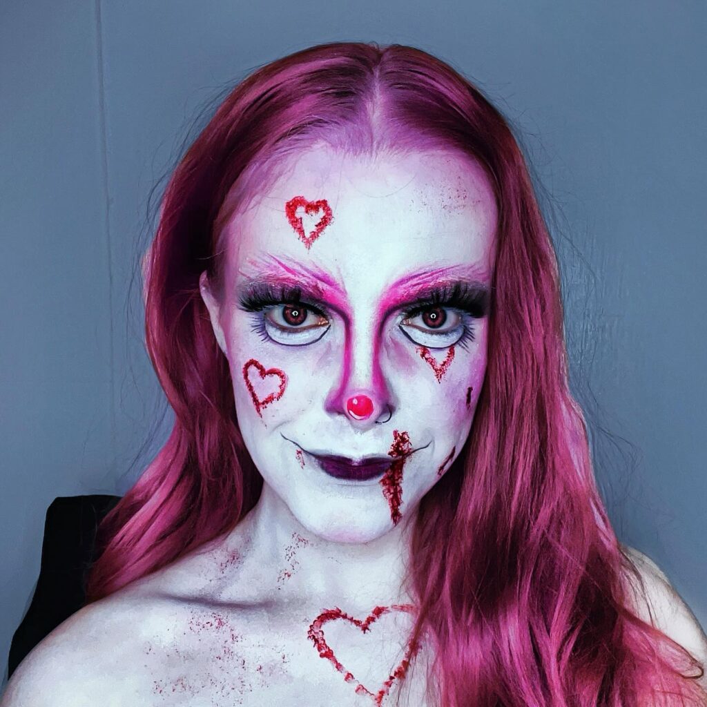 Person wearing heart-clown Halloween makeup with pink and white face paint, red hearts, dramatic eyelashes, and a red clown nose, complemented by pink hair.