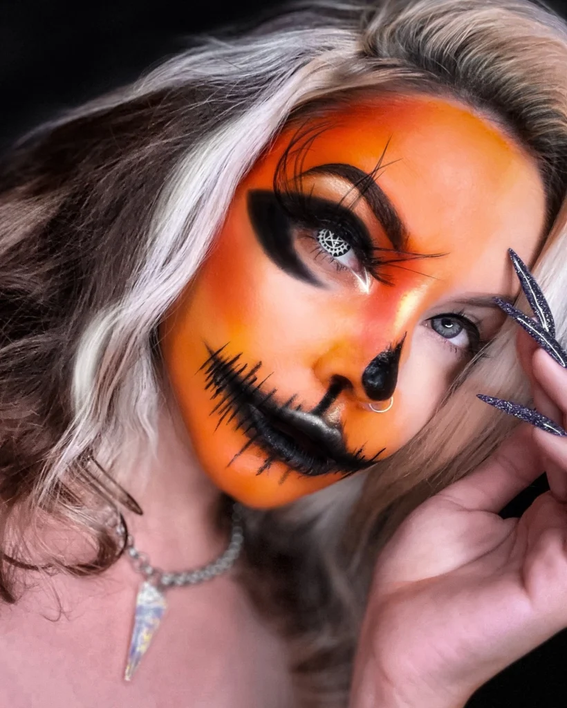 Close-up of a woman with Halloween makeup resembling a glowing pumpkin, featuring orange face paint, black dramatic eye makeup, stitched mouth detailing, and spider web contact lenses.