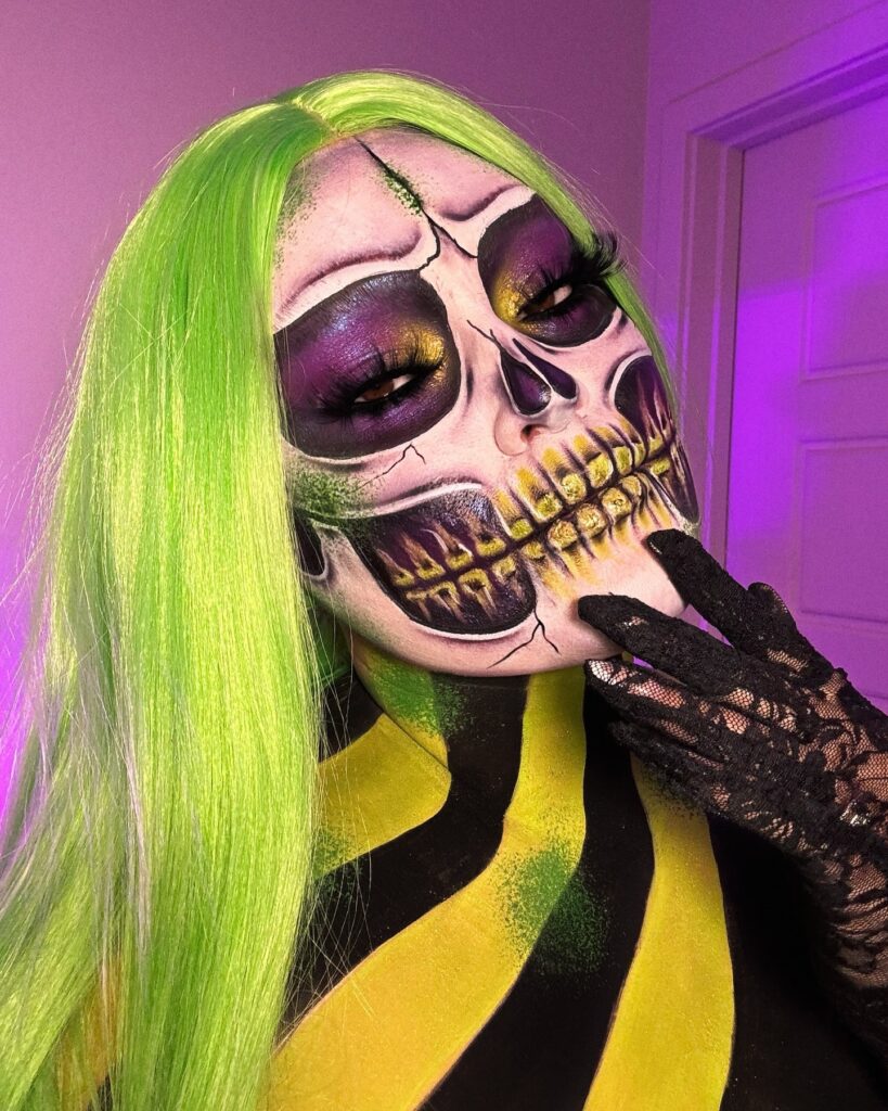 Person wearing neon green and purple skeleton-themed Halloween makeup with bright green hair, purple eyeshadow, and intricate skull contouring for a bold, electrifying look.