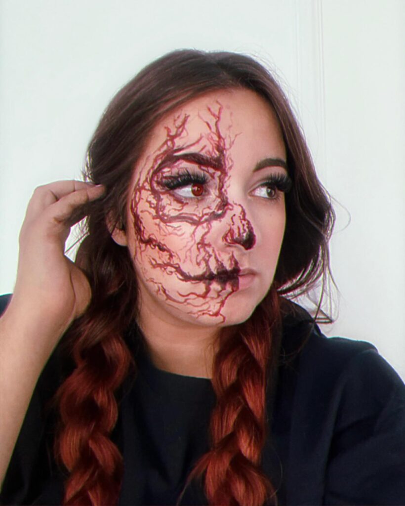 Person wearing veiny Halloween makeup with bloodshot veins and dark shading on half of the face, complemented by long lashes and braided hair for a creepy and unsettling effect.