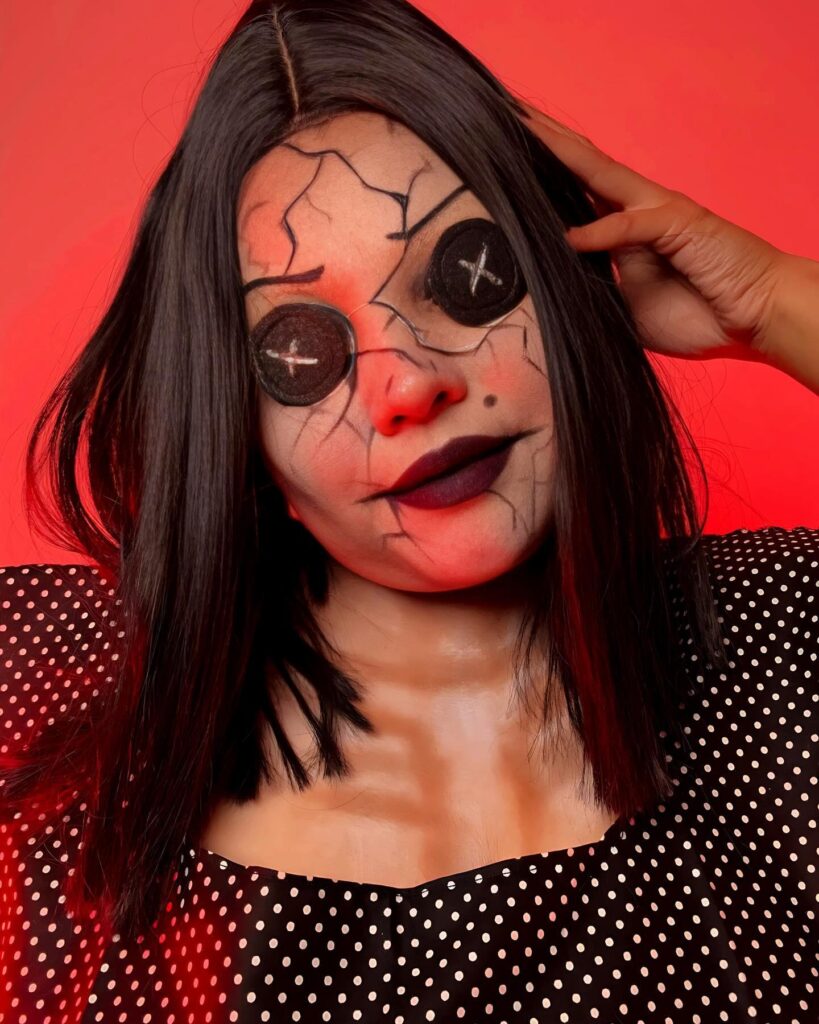 Person with black button eyes, cracked face makeup, and dark lipstick, wearing a polka-dotted top, against a red background for a haunting doll-like appearance.