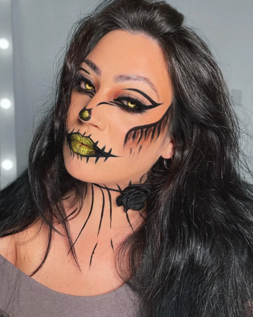 Close-up of a woman with Halloween makeup featuring a glamorous scarecrow design, including black stitching details, dramatic winged eyeliner, and metallic gold lips. The look is both eerie and stylish, blending spooky and glamorous elements seamlessly.