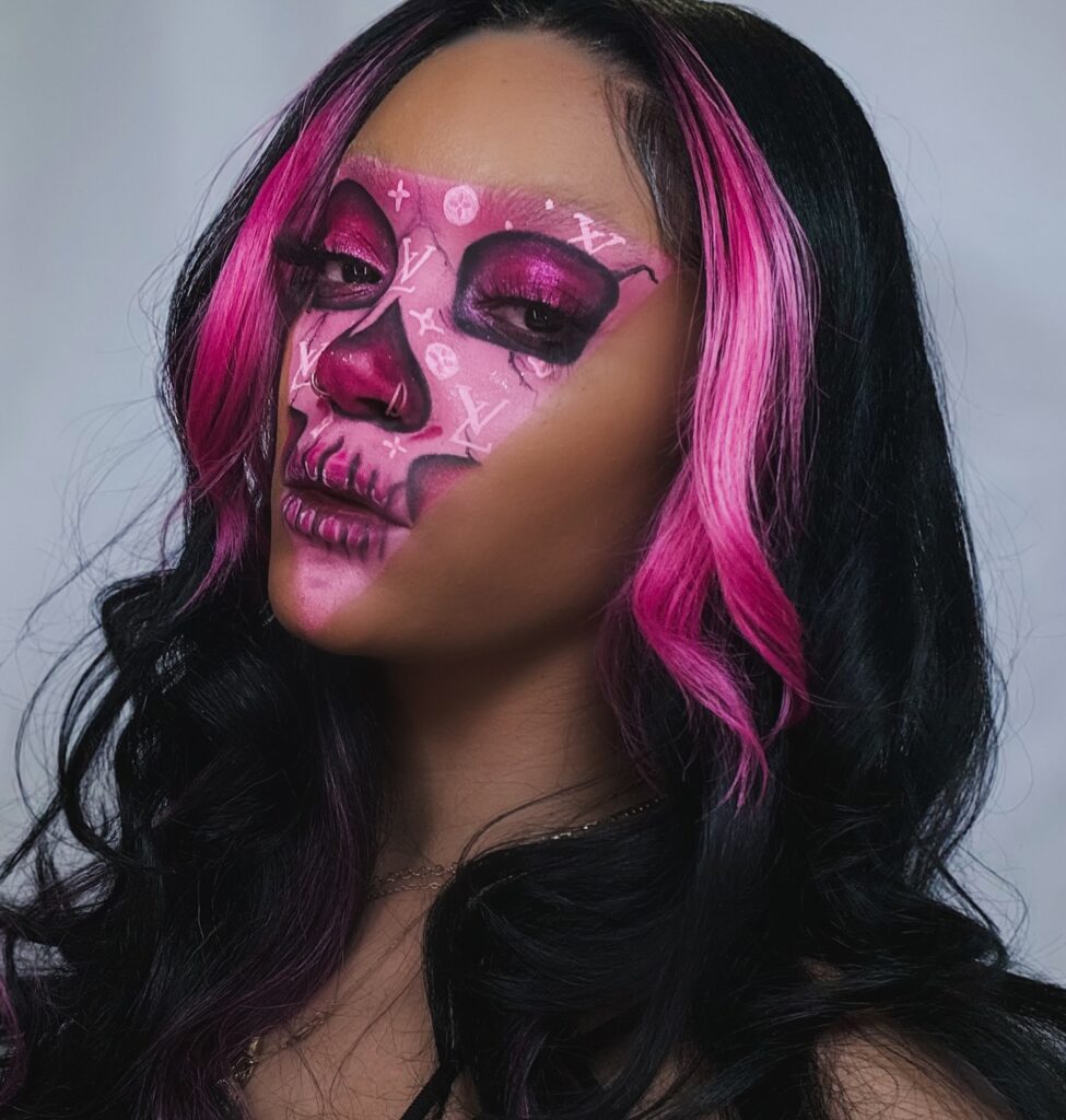 Person wearing luxury-inspired Halloween makeup with a pink skull design, white luxury logos, bold pink eyeshadow, and matching pink highlights in black hair for a chic, spooky look.