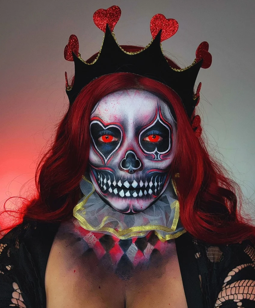 Person wearing Queen of Hearts-inspired skull makeup with red and black skeletal details, heart-shaped eye accents, red contacts, and a heart-topped crown for a regal and haunting Halloween look.