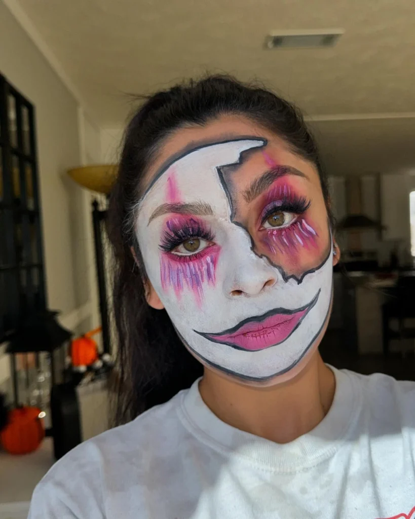 Close-up of a woman with Halloween makeup featuring a cracked doll face design, accented by vibrant pink eye makeup and soft pink lips. The cracked porcelain effect adds an eerie touch to the otherwise vibrant look.