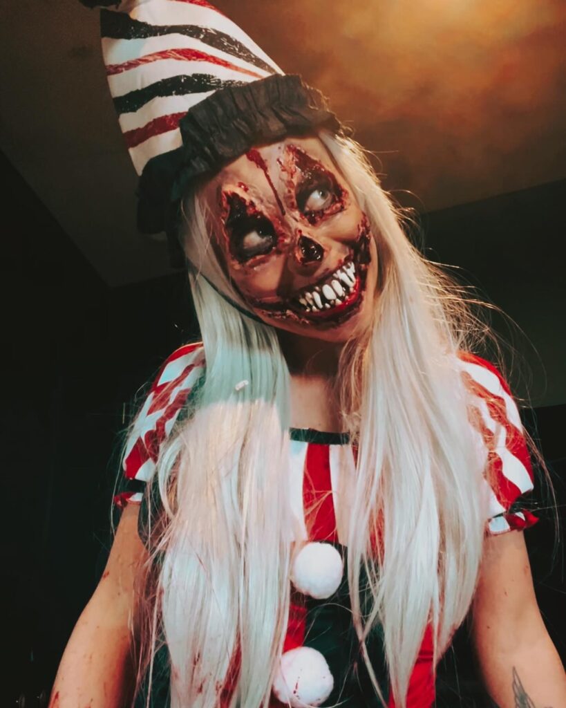 Person wearing terrifying circus clown Halloween makeup with bloodied eyes, sharp teeth, a striped hat, long white hair, and a red and white circus outfit.