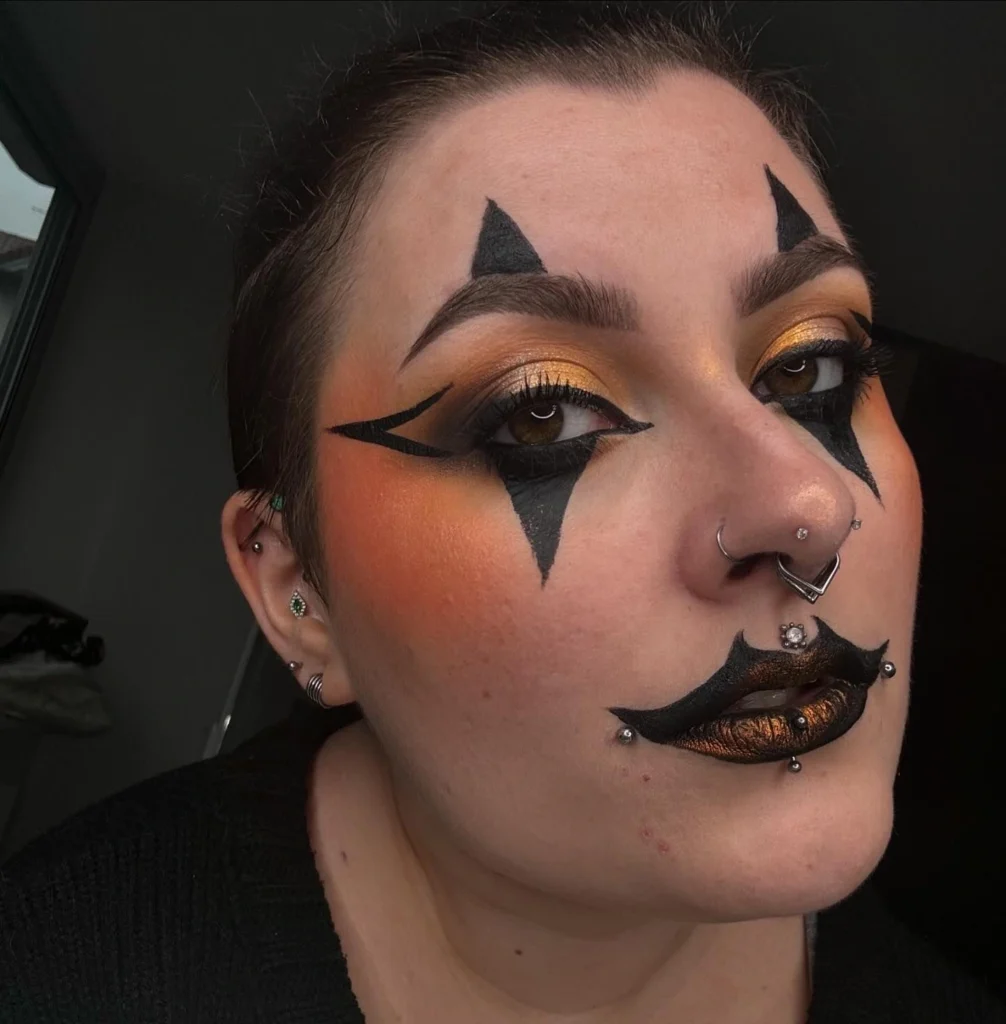 Close-up of a woman wearing gothic clown-inspired Halloween makeup with black triangles around the eyes, golden eyeshadow, and black lips highlighted with metallic gold.