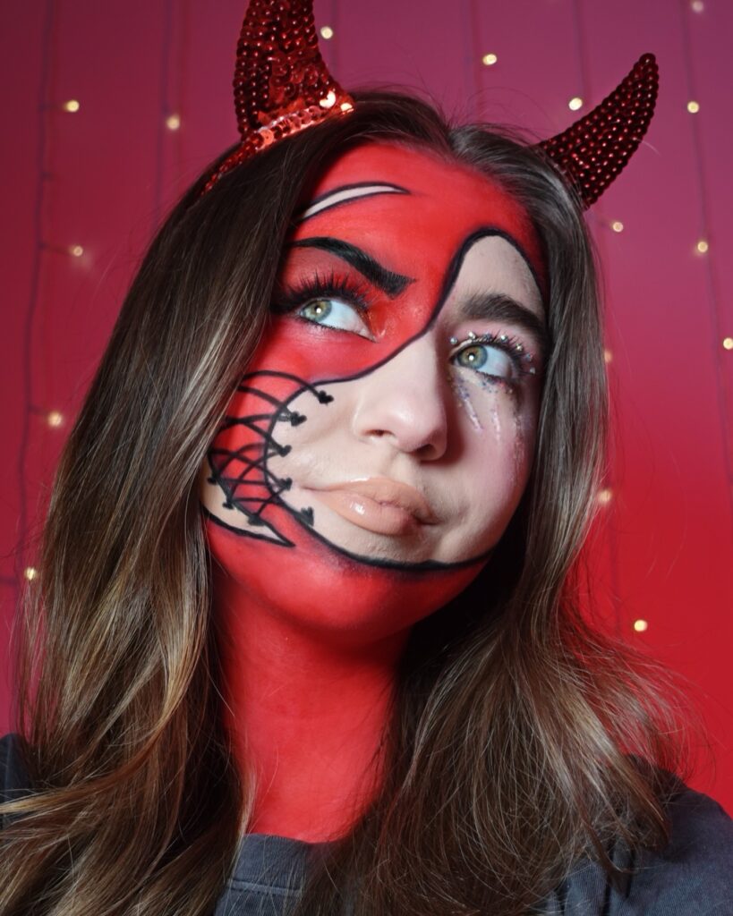 Person wearing bold devil-inspired face makeup with a red base, black detailing around the eyes and mouth, and glossy nude lips. The look is accentuated with sequined devil horns and a backdrop of warm glowing lights.