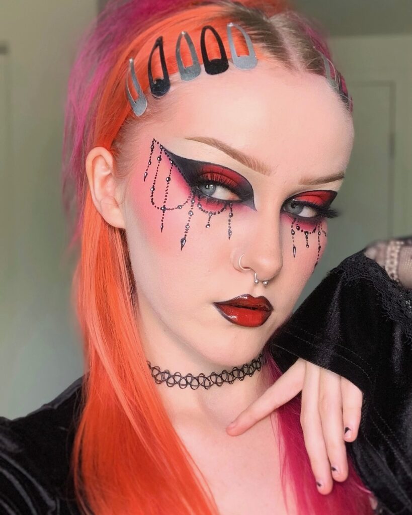 Person wearing edgy gothic Halloween makeup with bold red and black eyeshadow, winged eyeliner, black beaded chain designs under the eyes, glossy black and red lips, and orange and pink hair.