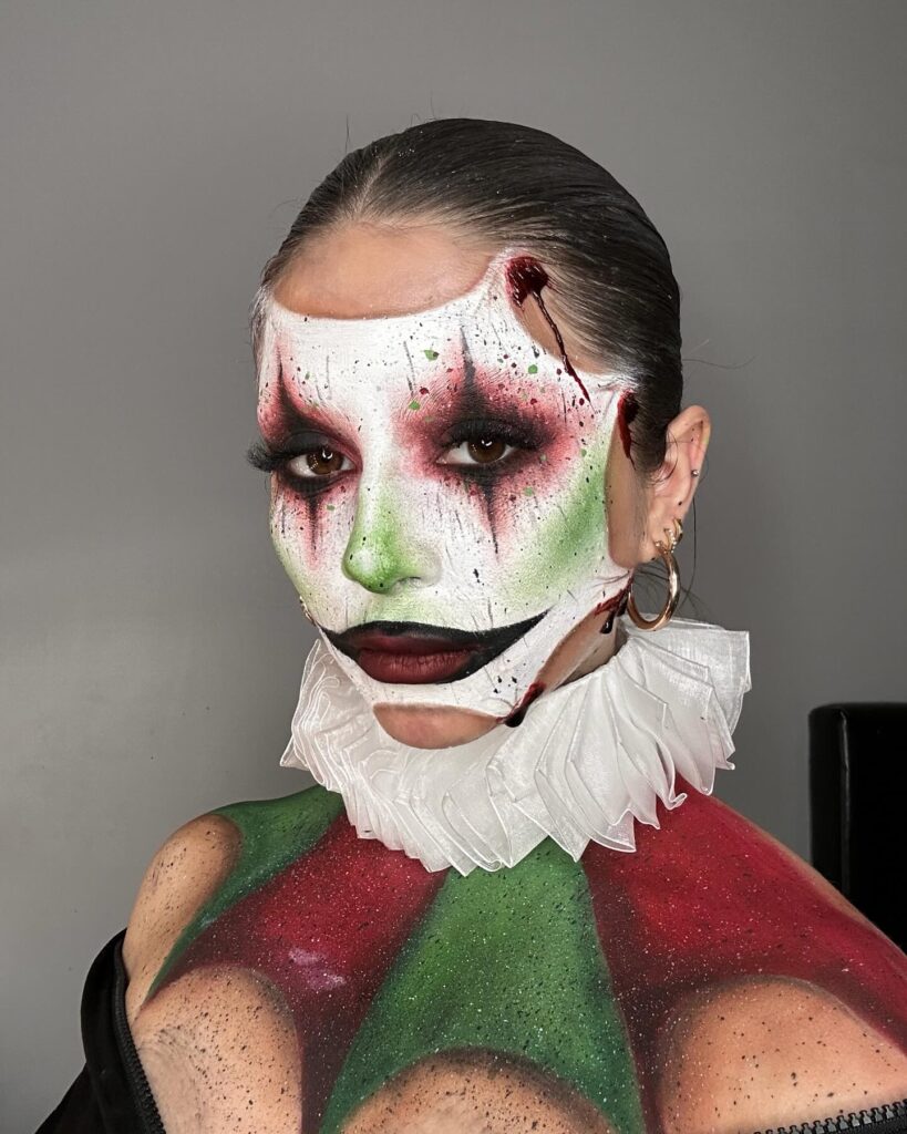 Person wearing Joker-inspired Halloween makeup with a white face, green and red shading, blood splatters, dramatic eyeshadow, and a sharp smile design, finished with a ruffled collar.