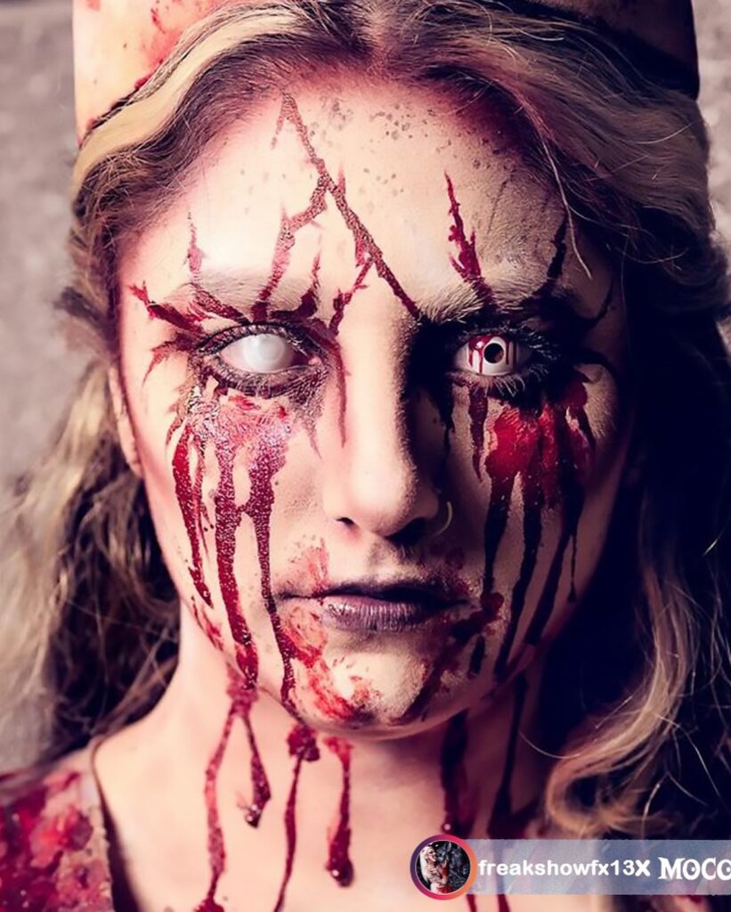 Person wearing bloody horror Halloween makeup with red veins, dripping blood, white contact lenses, and a pale complexion for a gory and terrifying look.