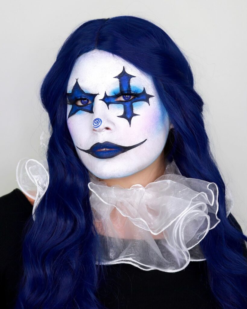 Person wearing blue clown Halloween makeup with black star-shaped eye accents, blue lips, a swirling blue nose, and a matching blue wig with a translucent ruffled collar for a haunting clown look.