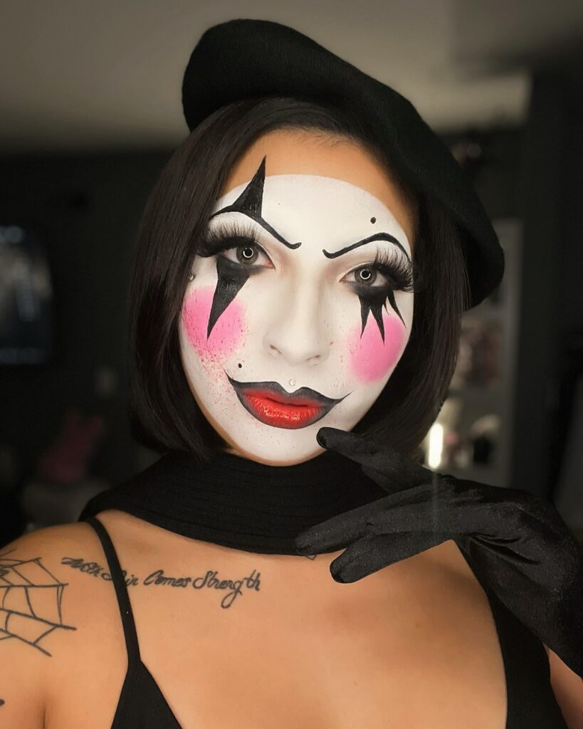 Person wearing classic mime Halloween makeup with white face paint, black tear-shaped accents, pink blush, glossy red lips, black beret, and black gloves for an elegant mime-inspired look.