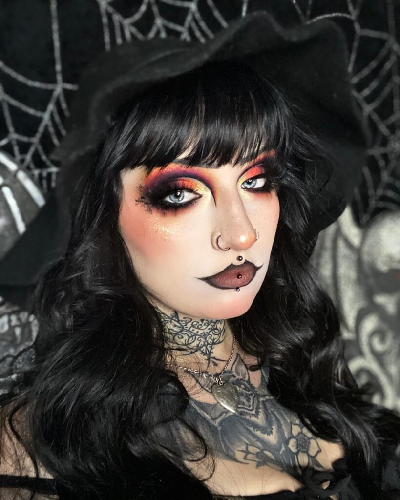 Person wearing glamorous gothic witch Halloween makeup with smokey red and orange eyeshadow, dark lips, black wavy hair, a witch hat, and intricate neck tattoos for a mysterious and enchanting look.