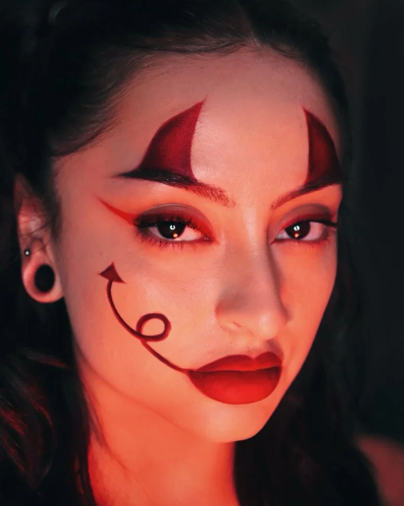 Close-up of a woman with devil-inspired Halloween makeup, featuring red horns painted on her forehead, red eyeshadow, and a curled devil tail design on her cheek.