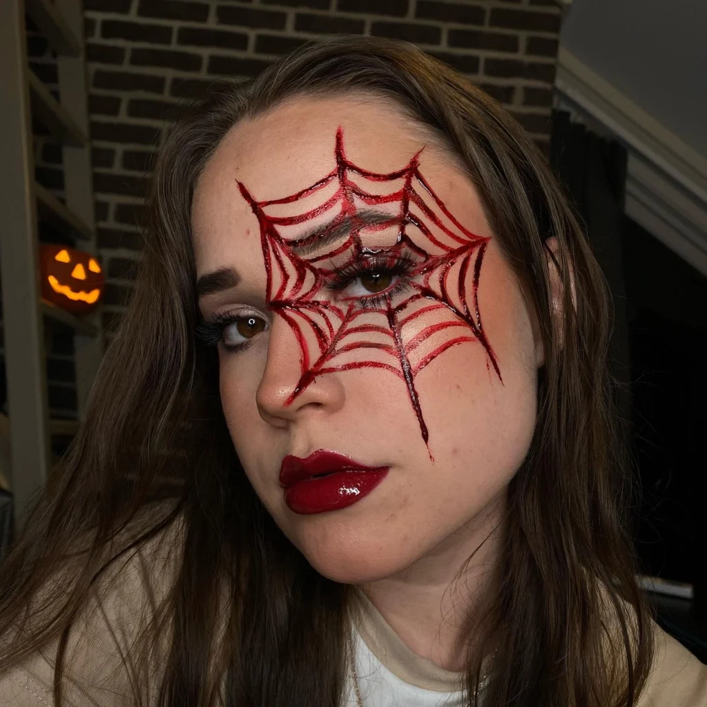 Close-up of a woman with Halloween makeup featuring a blood-red spider web design over one eye and glossy red lips, set against a background with a lit pumpkin decoration.