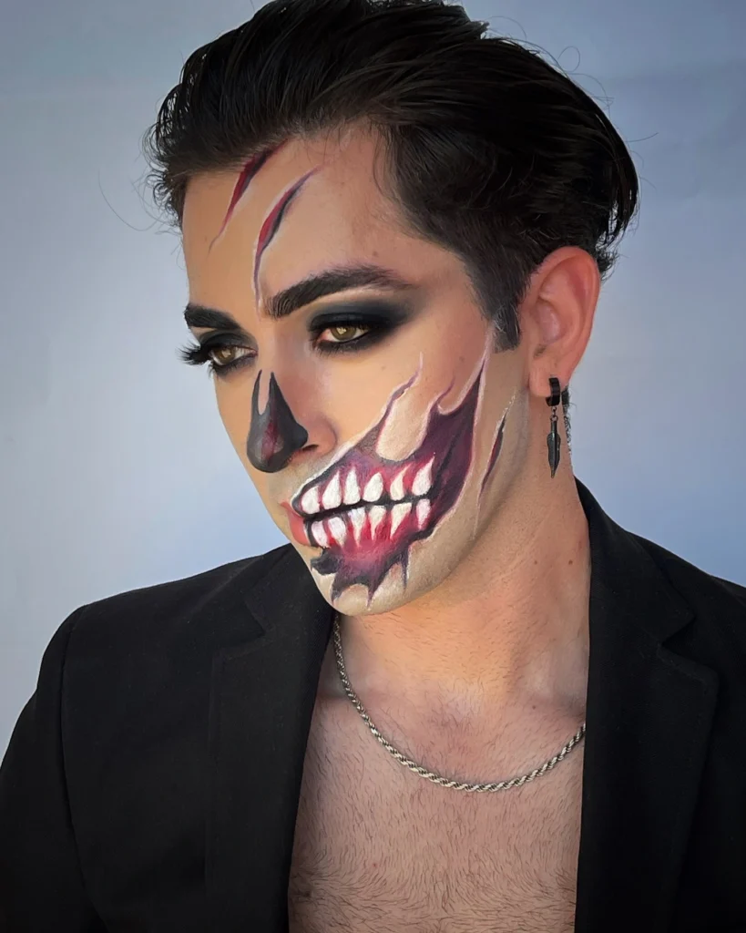 Close-up of a man with Halloween makeup featuring a skull demon design, including jagged teeth, hollow black eyes, and deep red facial scars. The makeup is highly detailed and creates a frightening, intense appearance.