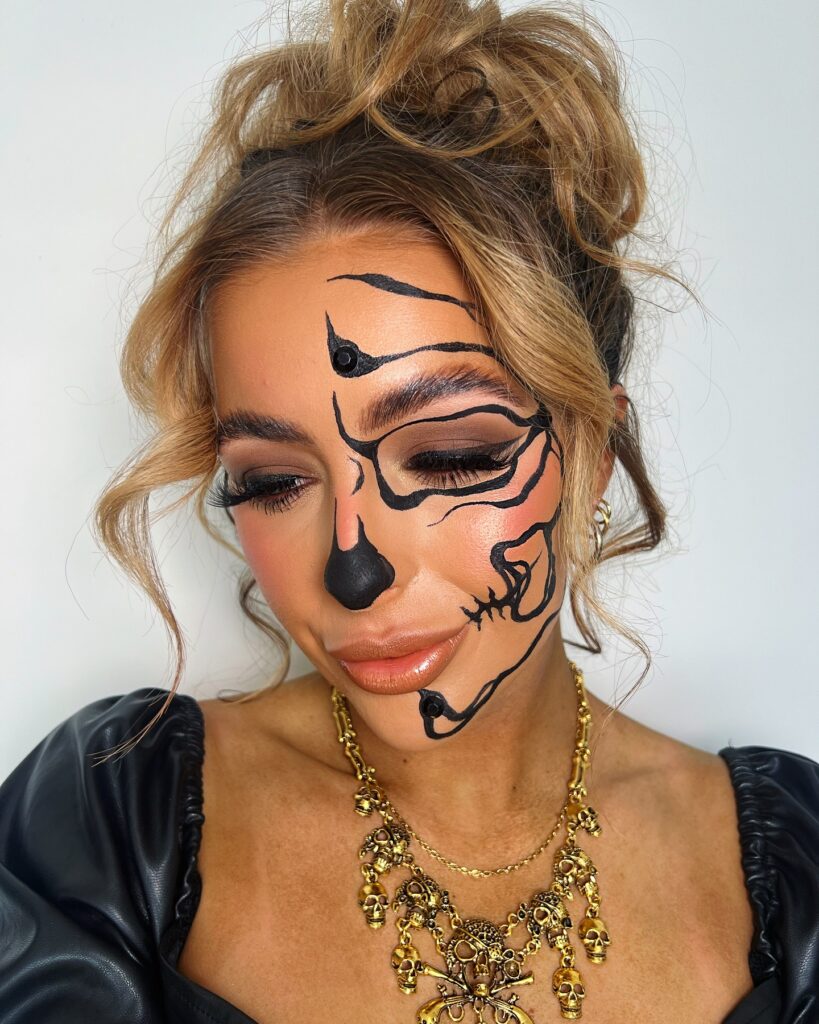 Person wearing minimalist skull Halloween makeup with black lines outlining facial bones, soft smoky eye makeup, glossy lips, and a statement gold skull necklace for a refined and spooky look.