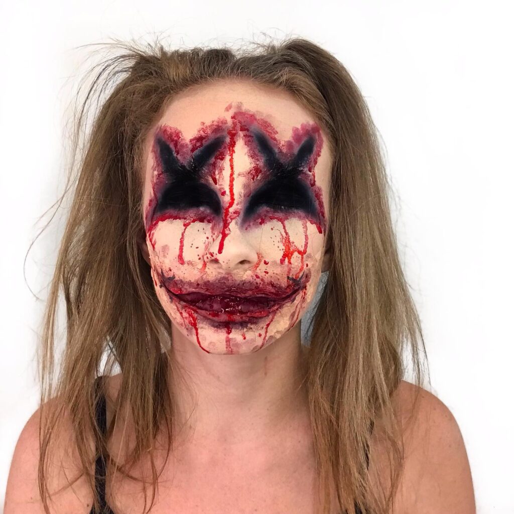 A person with a distressed clown makeup featuring deep black "X" eyes and a bloody, exaggerated smile, with blood dripping down the face and disheveled hair.