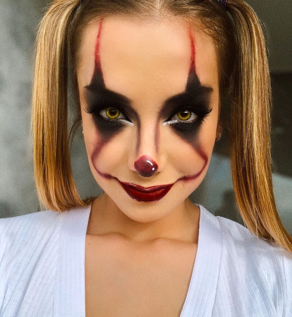 A person with dramatic smokey clown makeup, featuring dark black shading around the eyes, red accents on the brows and lips, and a shiny red nose, paired with sleek pigtails.