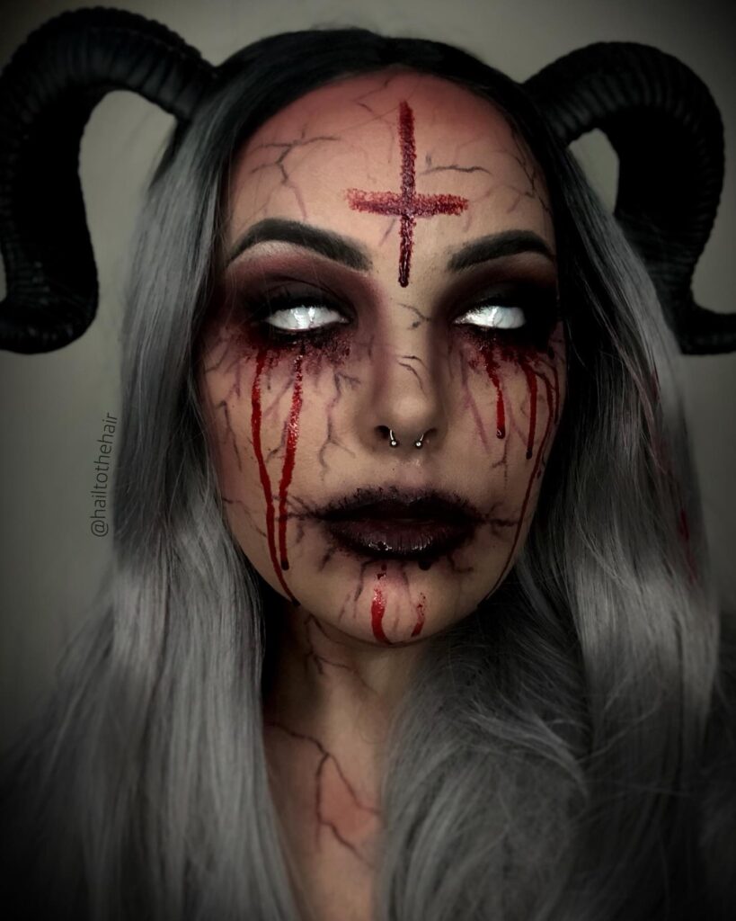 "A person with dark demon-inspired makeup featuring black eyeshadow, red tears, white eyes, and black horns with long gray hair.