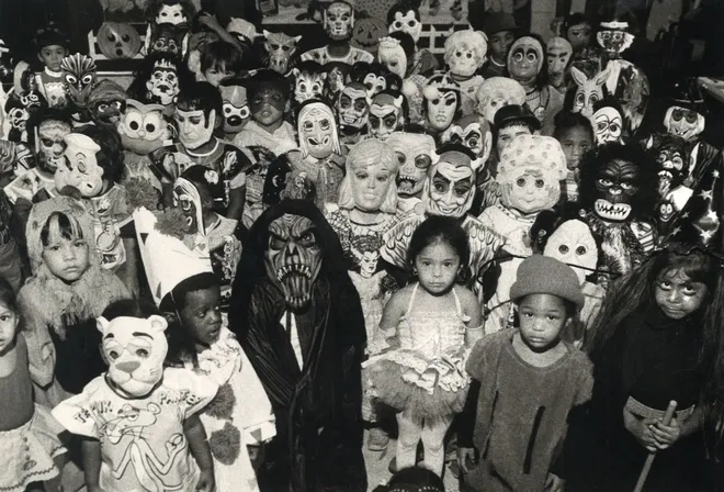 A black-and-white photograph from the 1980s showcasing a large group of children dressed in a variety of vintage Halloween costumes and masks. The costumes range from spooky to whimsical, capturing the eerie yet charming atmosphere of Halloween celebrations from decades past.