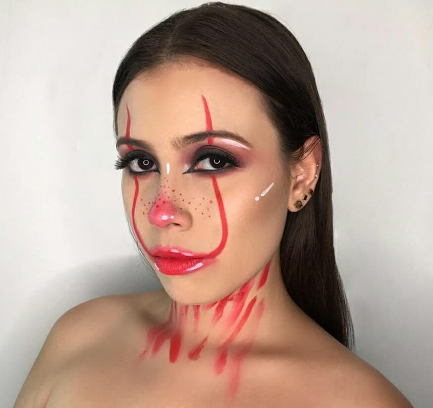 A person with minimalist clown makeup, featuring red lines and blush accents around the eyes, nose, and lips, with white highlights and a sleek hairstyle.