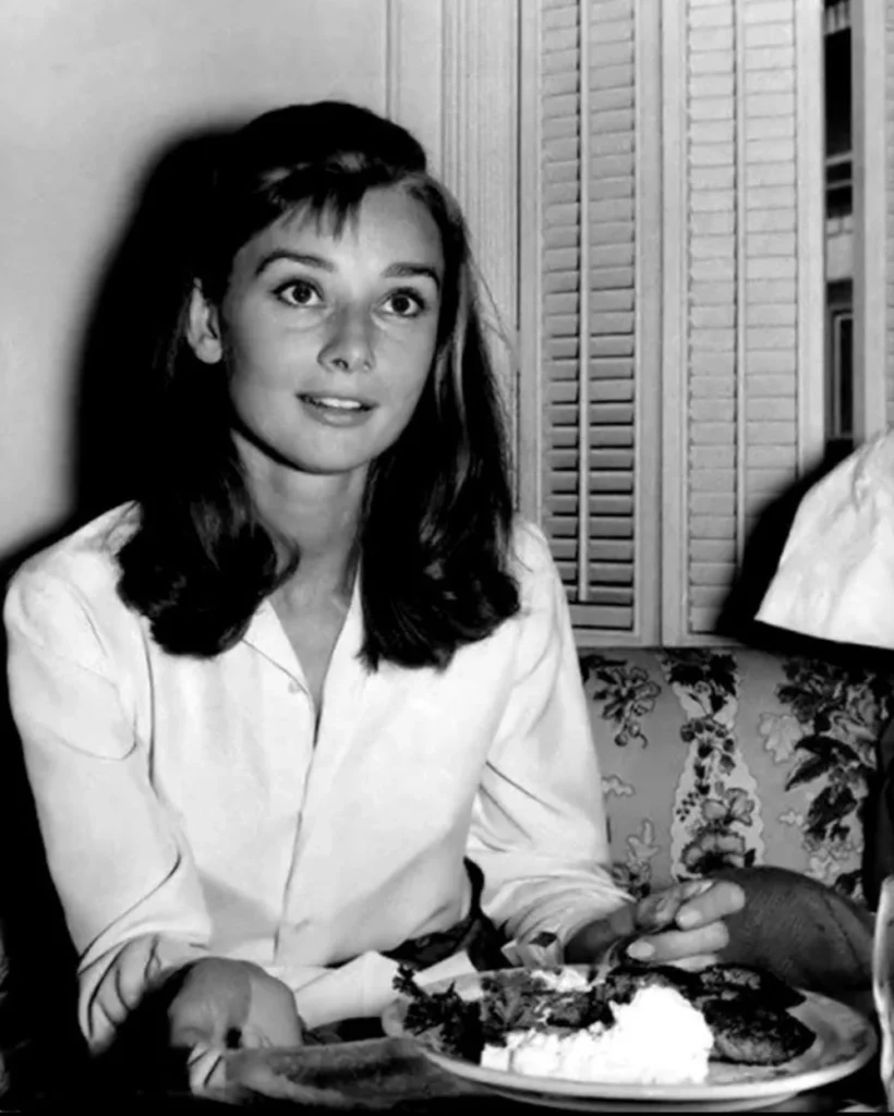 A black-and-white photograph of Audrey Hepburn, capturing her youthful elegance as she sits at a table with a plate of food, embodying the timeless grace and classic style she is renowned for.